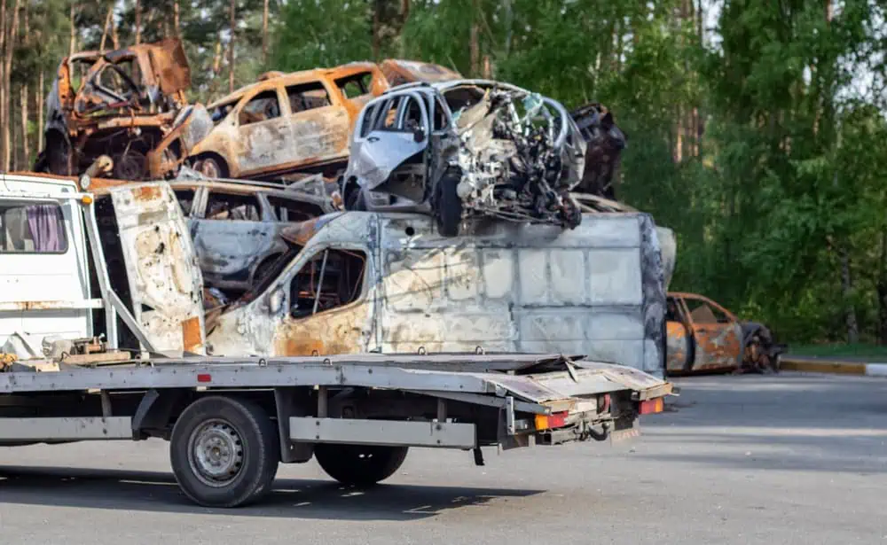 A flatbed tow truck is parked in front of a pile of rusted, burned-out vehicles stacked haphazardly, suggesting a scrapyard or accident aftermath. The scene implies a business where you can buy junk cars, with trees in the background enhancing this gritty tableau.