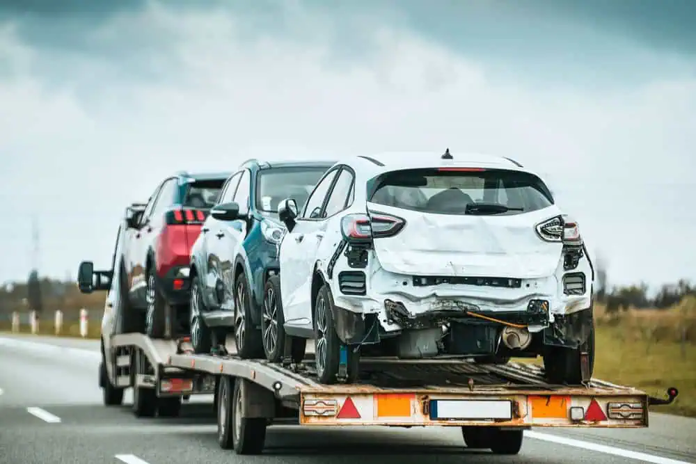 A tow truck, offering roadside assistance, carries damaged vehicles on a highway. The lead car has extensive rear damage, and the car behind is similarly affected. Overcast skies and blurred road details suggest movement and a rural setting.