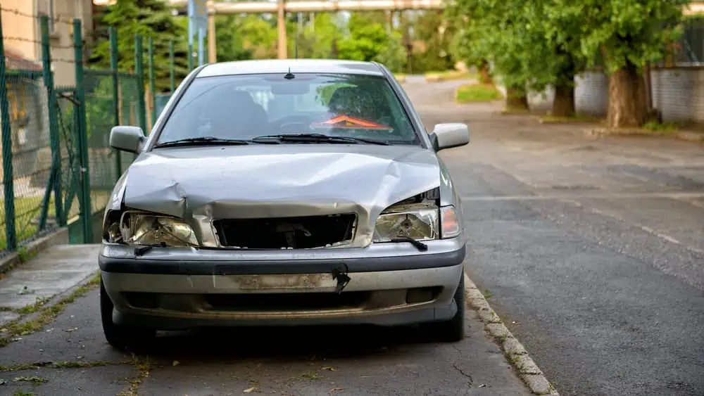 A silver car with a damaged front bumper and headlights is parked on the side of a quiet street, awaiting roadside assistance. The background displays green trees and nearby buildings.
