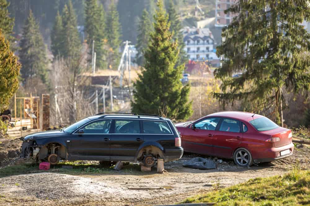 Two parked cars in a grassy area near trees; the black car on the left, typical of junk cars for cash, is missing its wheels and elevated on a jack. The red car on the right remains intact, with buildings visible in the background.