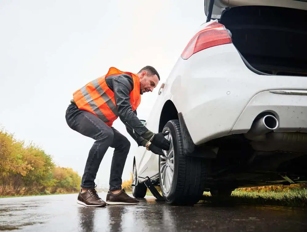 A man in a reflective orange vest kneels by the rear tire of a white car, offering roadside assistance on a rainy road. The car's trunk is open, and nearby trees boast autumn foliage. The wet road glistens from the rain, creating a challenging yet scenic backdrop for his diligent work.