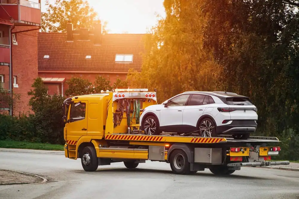 A yellow tow truck, offering roadside assistance, transports a white SUV on its flatbed along a quiet street. The backdrop features red brick buildings and lush, green trees. Sunlight casts a warm glow over the scene.
