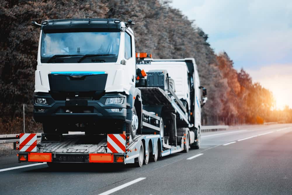 A car carrier truck is transporting a large white vehicle along a highway, perhaps one destined for new beginnings as part of our Junk Cars for Cash program. The road is lined with trees, and the sky is overcast. The truck and vehicle are prominent in the foreground.