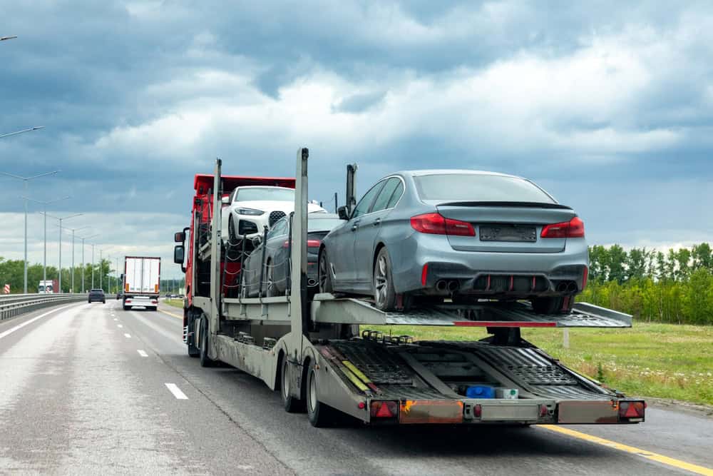 A car carrier truck, possibly offering roadside assistance, transports several vehicles, including a silver sedan on the upper deck, along a highway under a cloudy sky. In the background, greenery lines the road and a white truck follows.
