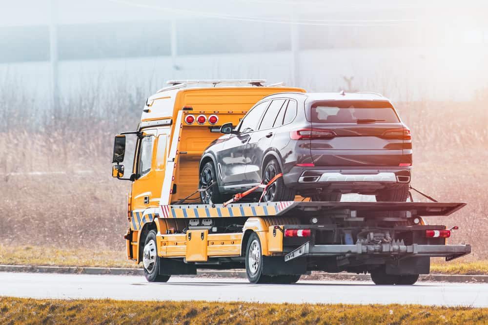 A tow truck, offering roadside assistance, carries a dark grey SUV on its flatbed along a road. The background reveals a grassy area and an industrial building, all captured on a sunny day.