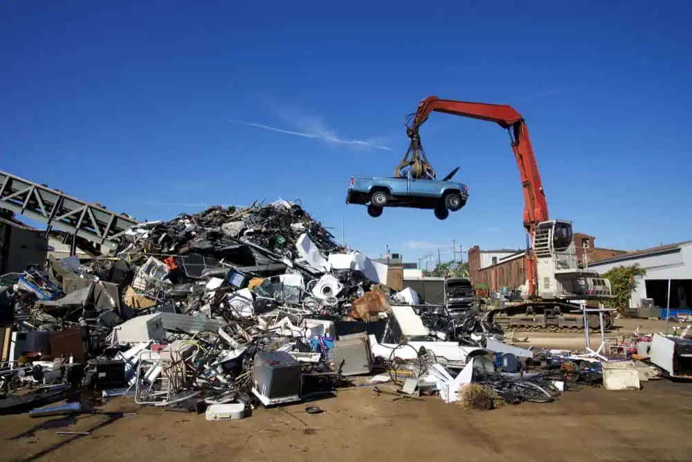 A pile of assorted scrap metal and electronic waste is seen under a clear blue sky. An excavator with an orange arm hoists an old blue car onto the heap in this industrial scrapyard, where they buy junk cars for cash, blending ecological responsibility with opportunity.