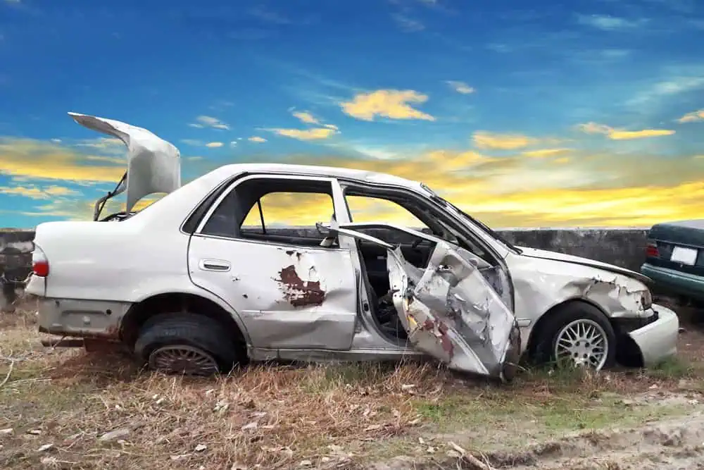 A heavily damaged white car with a crushed side and open doors sits abandoned on a grassy area. The trunk is ajar, the tires are flat, and it looks ready to sell your junk car. A vibrant sunset sky creates a stark contrast to the wreckage.