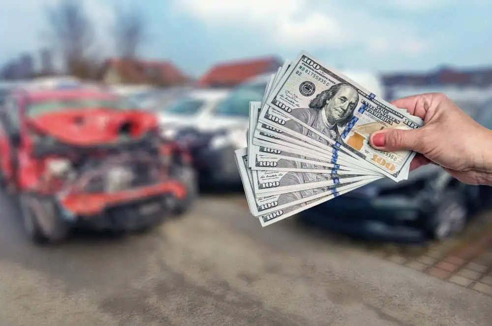 A hand holds several hundred-dollar bills with a background of a blurred, damaged red car and other vehicles in an outdoor setting, suggesting a transaction related to selling junk cars for cash or dealing with car damages.
