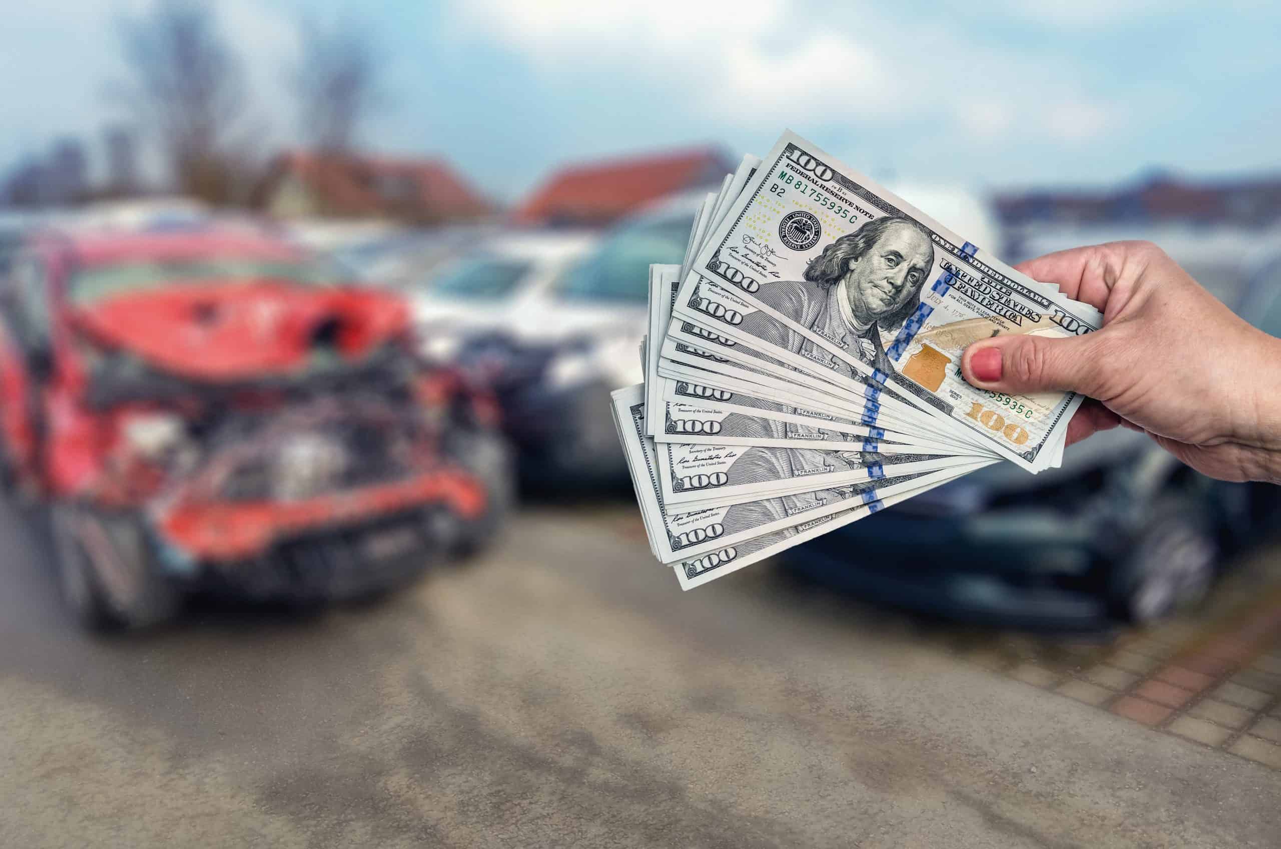 A hand holding a stack of 100-dollar bills is in focus against a blurred backdrop of a damaged red car in an outdoor parking lot. The overcast setting suggests it's time to sell your junk car and turn that clunker into cash.