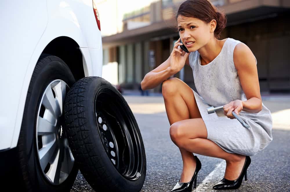 A woman crouches next to a car with a flat tire, wrench in hand, and talks on her phone. Wearing a light gray dress and black heels, she looks concerned in the city. Perhaps it’s time to sell your junk car for cash and buy something more reliable.