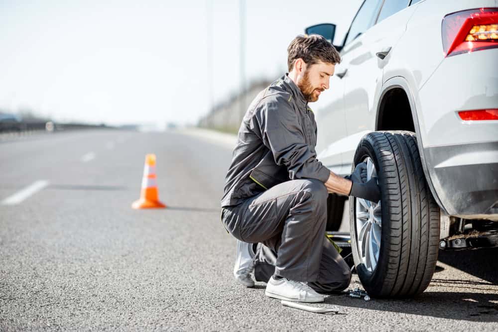 A man kneels on a highway beside a silver car, changing a tire with expertise akin to professional roadside assistance. Dressed in a gray uniform and gloves, he works diligently, an orange traffic cone placed nearby. The daylight highlights the empty road stretching into the distance.