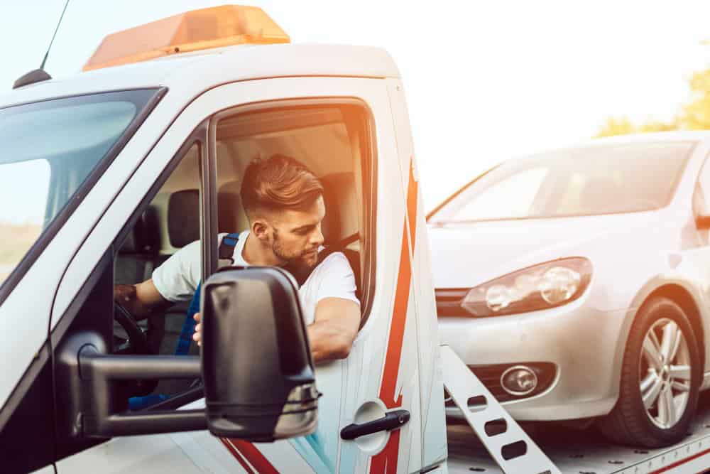 A tow truck driver with short hair sits in the driver's seat of a white tow truck adorned with red and blue stripes, offering roadside assistance. He looks back at a silver car hitched to the truck as the sun shines brightly in the background.