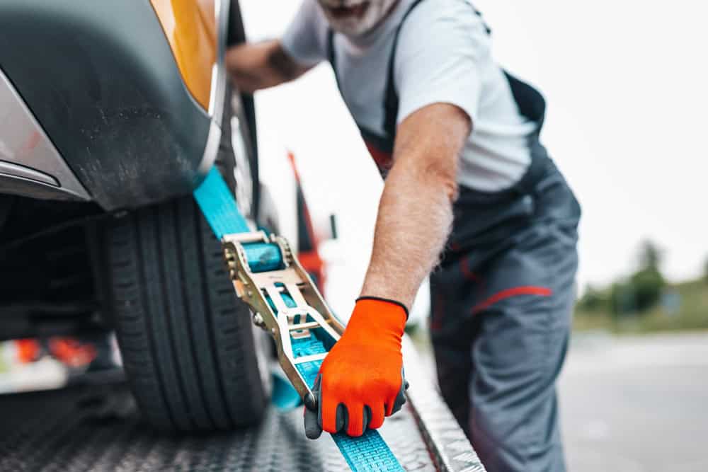 With orange gloves, a person secures a blue strap around the tire of a vehicle on a flatbed tow truck. The focus is on their hands and the ratchet strap mechanism, highlighting the meticulous roadside assistance process in progress.