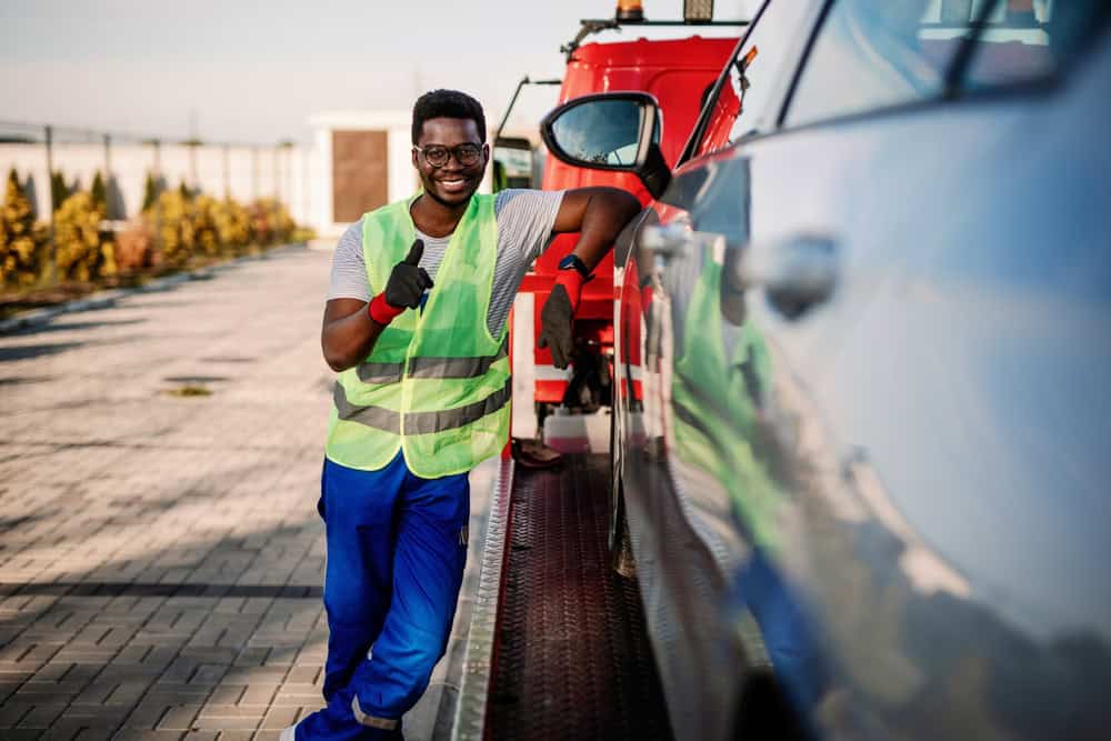 A worker in a high-visibility vest and blue pants smiles and gives a thumbs-up next to a parked tow truck with a reflective surface, suggesting successful roadside assistance or perhaps an opportunity to sell your junk car on this sunny day.