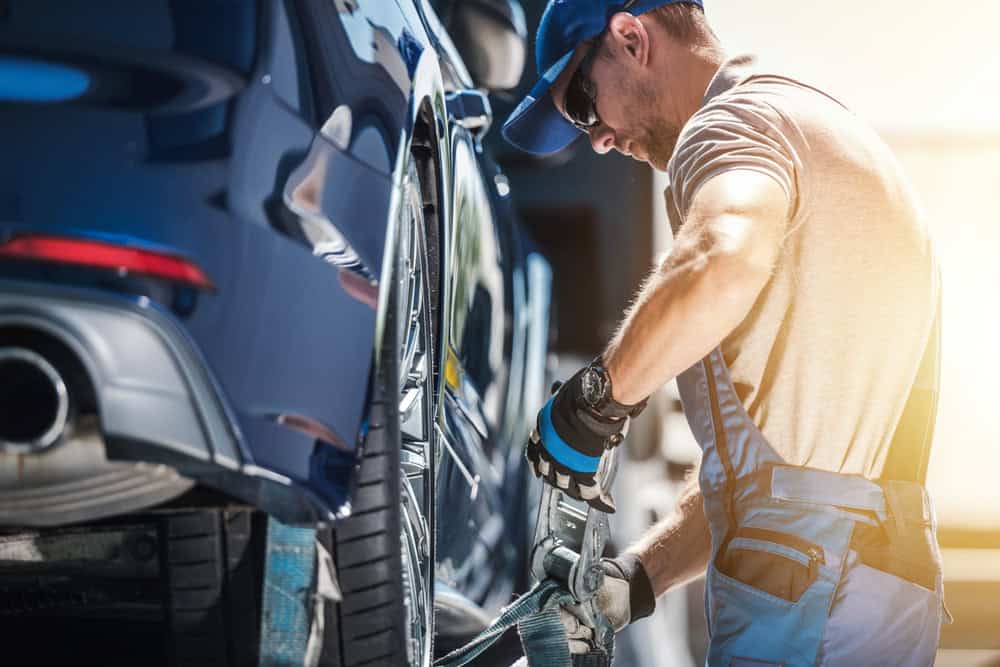 A mechanic in overalls and a cap secures a blue car onto a flatbed tow truck, part of his roadside assistance duties. The sun shines brightly, casting a warm glow on the scene as he ensures the vehicle is tightly fastened.