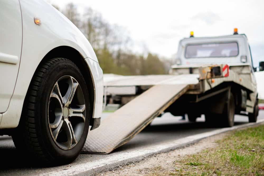 A white car, possibly a candidate for "sell your junk car," is parked on the roadside with its front wheel near a towing truck's ramp, ready for loading. Blurred trees form the backdrop of this roadside scene.