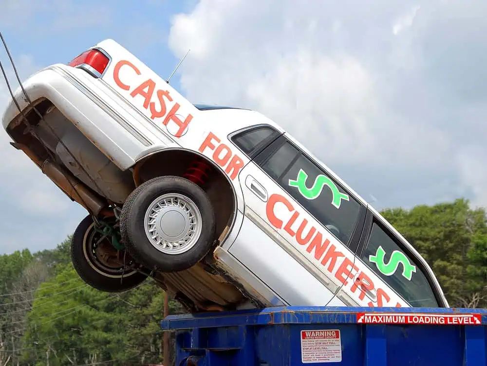 A white car with "Cash for Clunkers" painted in red is being lifted into a blue container, showcasing how easy it is to sell your junk car. Green dollar signs are drawn on the windows. Trees and a cloudy sky are in the background, framing this effortless handover.