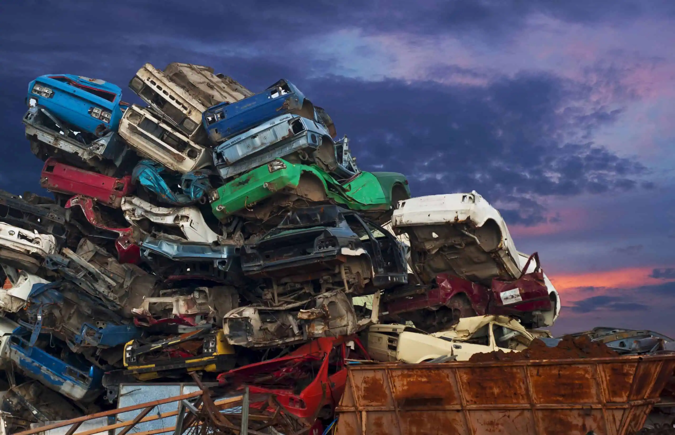 A large pile of crushed cars stacked in a scrapyard against a dramatic evening sky, waiting for buyers who specialize in junk cars. The colorful, rusted metal stands out against dark clouds with hints of sunset, inviting you to sell your junk car and clear the clutter.