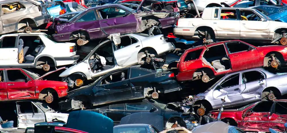 A large pile of damaged and stacked cars of various colors, including red, white, and purple, fills the scrapyard. Many vehicles are missing wheels, windows, or other parts. This cluttered collection is a haven for those looking to buy junk cars or sell your junk car for cash.