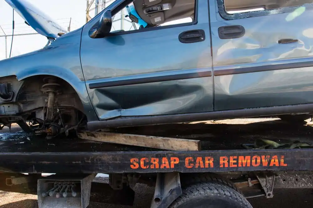 A damaged blue car, missing its front wheels, is loaded onto a truck labeled "Scrap Car Removal," ready for you to sell your junk car. The scene unfolds on a sunny day, right by a fence.