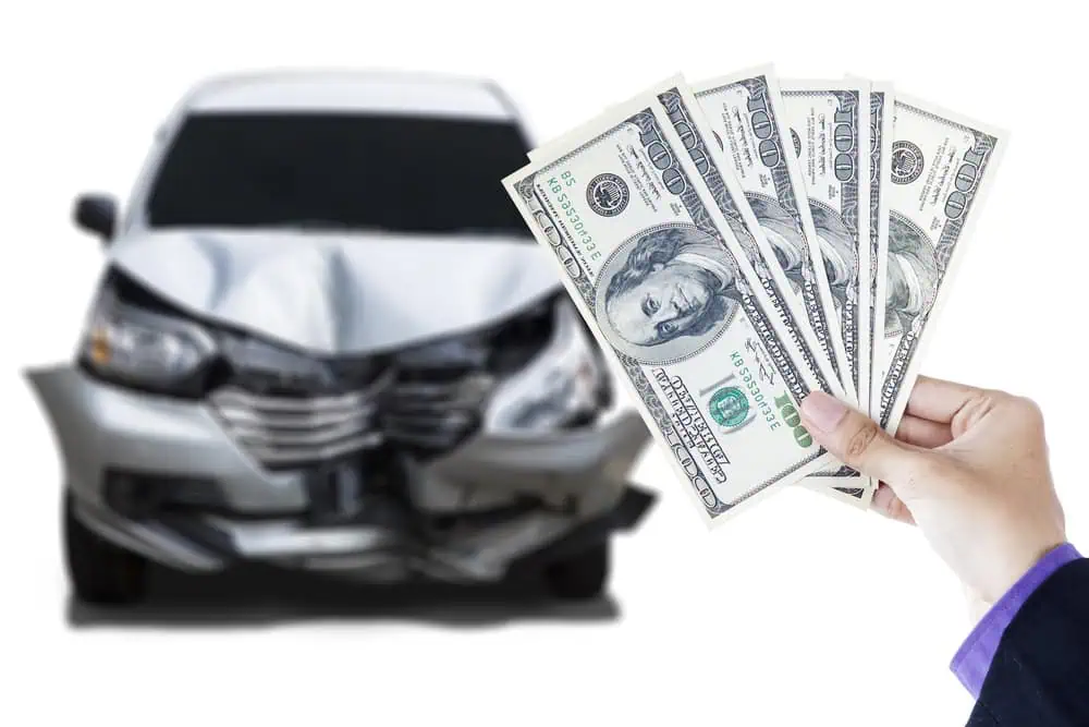 A hand holding several U.S. dollar bills is in focus, symbolizing financial compensation or costs related to car accidents, while a blurred silver car with a crumpled front bumper looms in the background&mdash;reminding us that selling your junk car could convert wreckage into cash.