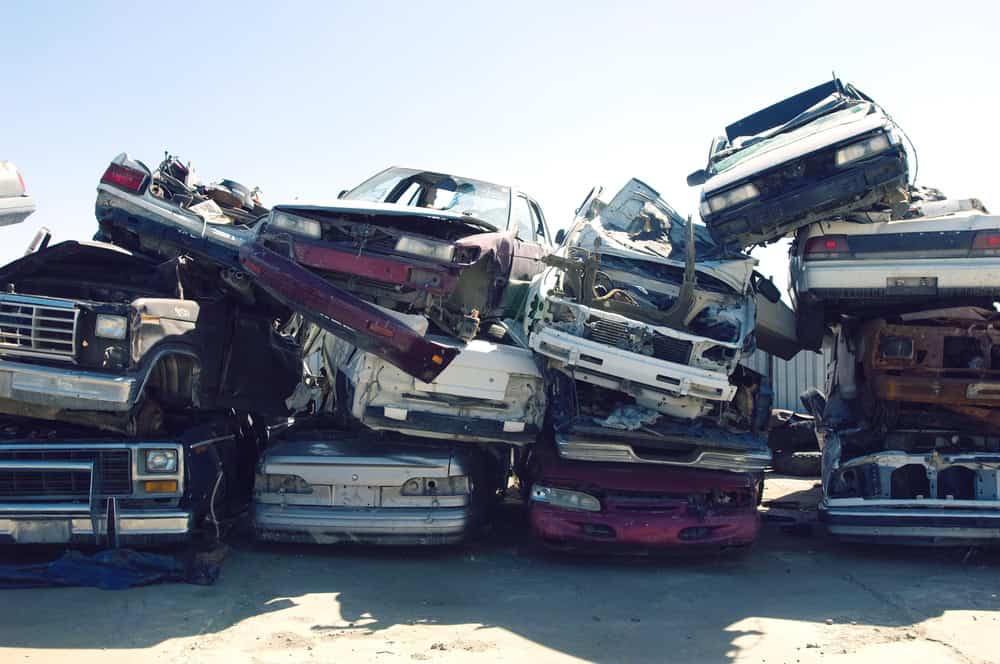 A pile of old, crushed cars stacked haphazardly in a junkyard under a clear blue sky. The vehicles are in various states of disrepair, with missing hoods and doors, showcasing rusted frames. Consider turning these eyesores into profit with our "Junk Cars for Cash" program today!